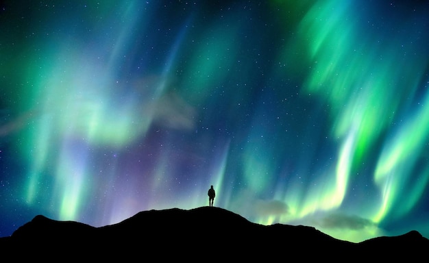 Aurora boreal brilhando sobre o caminhante de silhueta em pé na montanha no céu noturno