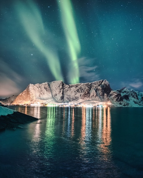 Aurora Borealis, Nordlichter über schneebedeckten Bergen mit leuchtendem Dorf in Hamnoy auf den Lofoten, Norwegen
