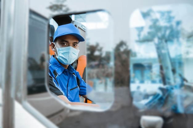 Aus dem Fenster eines männlichen Fahrers in Uniform gesehen und mit Maske in die Kamera schaut, während er im Bus sitzt sitting