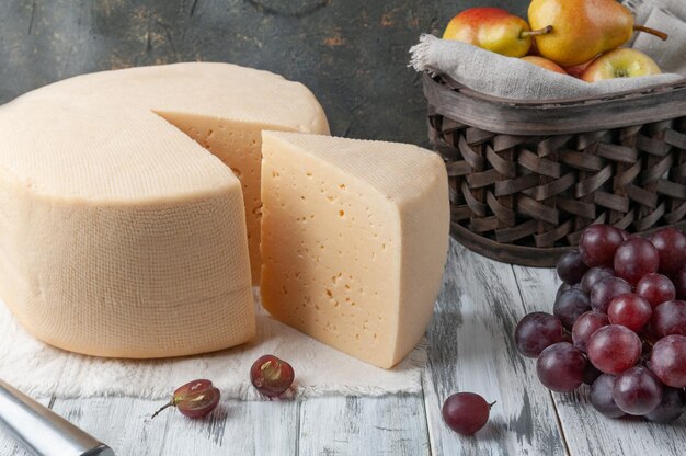 Aus dem runden Käsekopf wird ein dreieckiges Stück geschnitten. Käse liegt auf einem Leinentuch. In der Nähe ein Messer, Trauben und ein Obstkorb. Auf einem grauen hölzernen Hintergrund. Platz für ein Logo. Nahansicht.