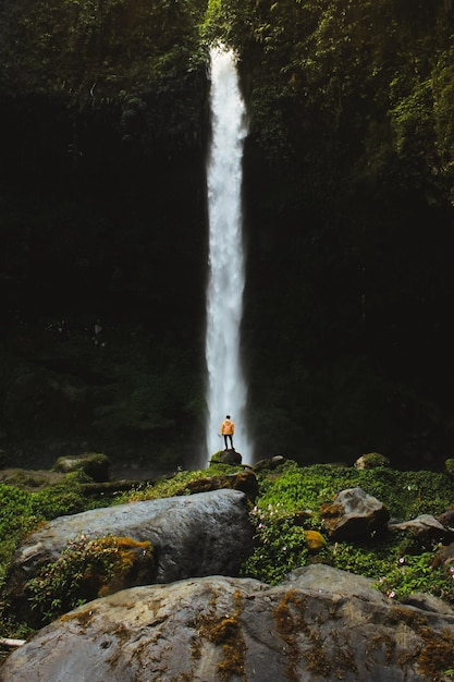 Aus mittlerer Entfernung sieht man einen Teenager, der am Wasserfall im Wald steht