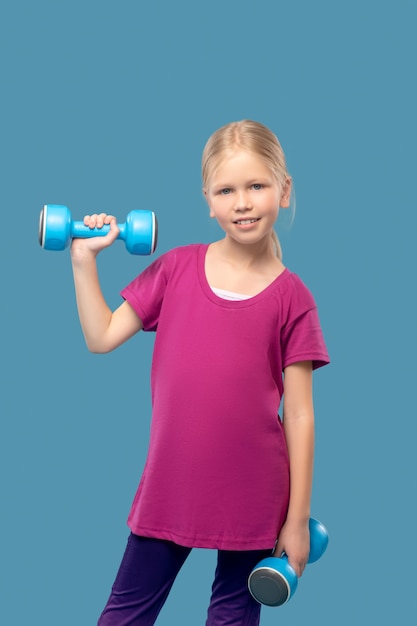 Ausbildung, Härten. Fröhliches Mädchen mit langen blonden Haaren im Sportbekleidungstraining mit Hanteln auf blauem Hintergrund im Studio