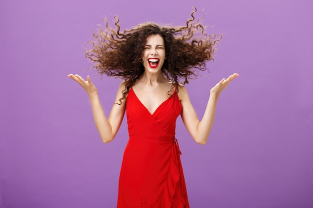 Ausdruck von innerer Wildheit. Porträt einer wilden und sorglosen, charmanten europäischen Frau in stilvollem rotem Abendkleid, die lockiges Haar mit erhobenen Händen hochschnippt und sich während der Party laut lacht.