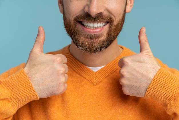 Ausgeschnittene Ansicht des erfolgreichen positiven bärtigen Mannes, der große Finger in die Kamera mit einem zahnigen Lächeln zeigt Studioaufnahme im Innenbereich isoliert auf blauem Hintergrund