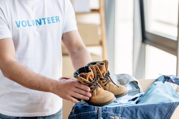 Foto ausgeschnittene ansicht eines freiwilligen, der kinderschuhe hält, während er neben einem karton mit jeans steht