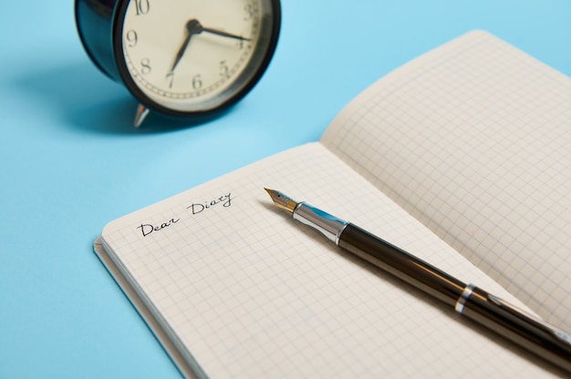 Ausgeschnittene Ansicht eines offenen Notizblocks mit dem Wort Liebes Tagebuch, Wecker und einem Tintenstift einzeln auf blauem Hintergrund mit Kopierraum