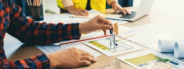 Foto ausgeschnittenes bild eines professionellen ingenieurteams, das an einem bauplan arbeitet, während sein kollege am laptop am besprechungstisch arbeitet, mit bauplänen und holzblöcken, die überall verstreut sind. nahaufnahme der abgrenzung