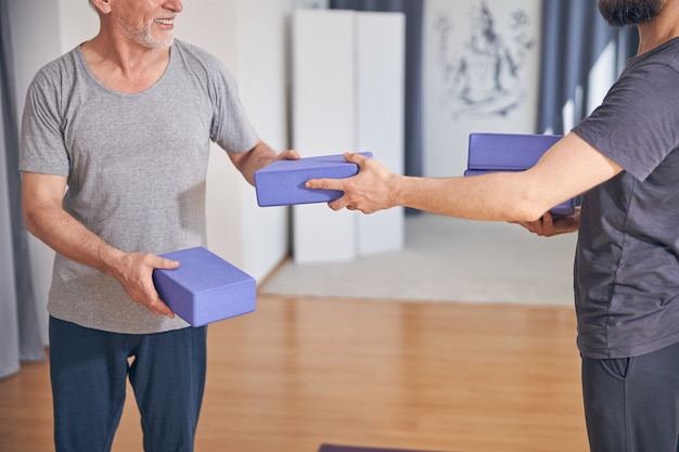 Ausgeschnittenes Foto eines lächelnden reifen Mannes, der die Yogablöcke von einem bärtigen Trainer nimmt taking