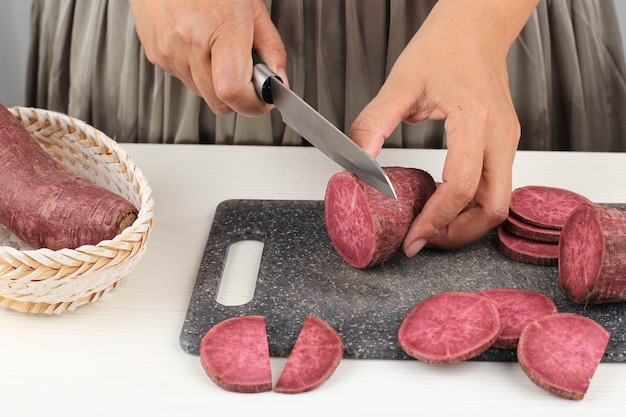 Ausgewählter Fokus A Woman Slice Cut japanische lila Süßkartoffel mit Messer in der Küche, weißer Hintergrund, Küchenprozesskonzept