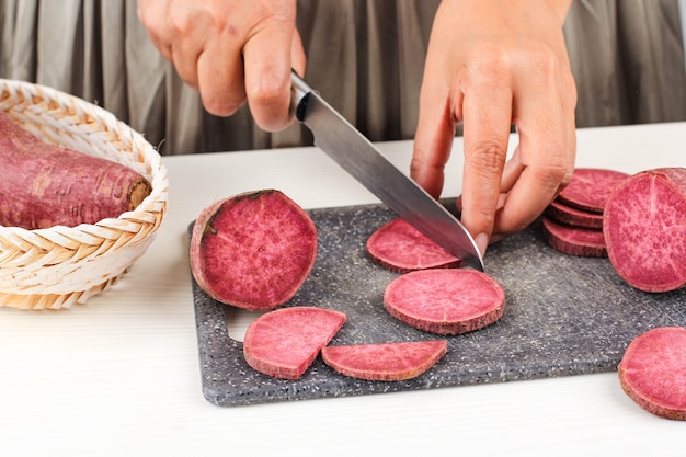 Ausgewählter Fokus A Woman Slice Cut japanische lila Süßkartoffel mit Messer in der Küche, weißer Hintergrund, Küchenprozesskonzept