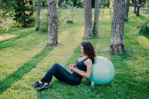 Außenaufnahme einer entspannten brünetten Frau, die in der Nähe eines Fitnessballs sitzt, benutzt ein modernes Mobiltelefon, hört Musik über Kopfhörer, posiert auf grünem Gras mit einer Flasche Wasser, trägt Sportkleidung und Turnschuhe