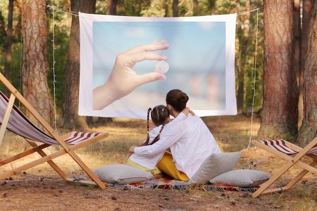 Außenaufnahme einer glücklichen positiven Mutter und Tochter, die im Wald sitzen und Filme oder Fotos auf der Projektorfamilie ansehen, die Zeit miteinander verbringen und die schöne Natur genießen