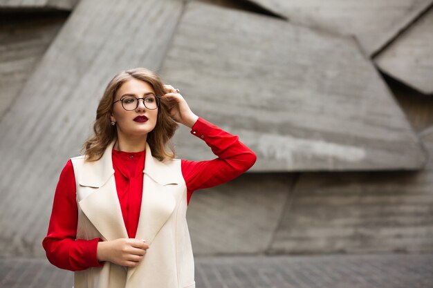 Außenaufnahme einer schönen blonden Frau mit roten Lippen, die weiße lange Weste und Brille trägt. Platz für Text