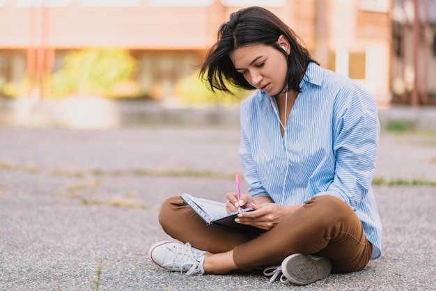 Außenaufnahme einer schönen jungen kreativen Autorin schreibt einige Ideen in den Notizblock. Verwendet Smartphone für die Kommunikation. Hören Sie die Lieblingsmusik auf der Straße. Brünette Studentin bereitet Prüfungen vor