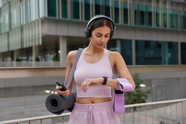Außenaufnahme einer sportlich fitten Frau, die mit Sportausrüstung posiert, überprüft die Zeit auf der Smartwatch und nutzt das Smartphone, um Fitnessergebnisse zu verfolgen, die in Sportswear-Posen im Freien vor urbanem Hintergrund gekleidet sind