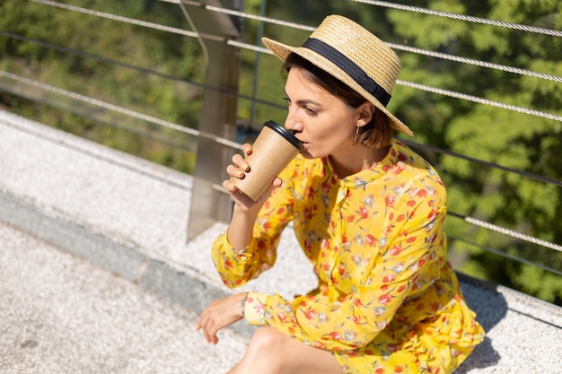 Außenporträt der Frau im gelben Sommerkleid und im Hut mit der Tasse Kaffee, die Sonne genießt