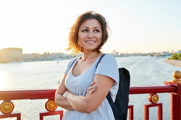 Außenporträt der reifen schönen glücklichen Frau mit verschränkten Armen, weibliches Gehen mit Rucksack auf Brücke am Sommertag, goldene Stunde