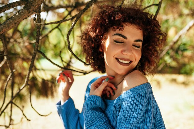 Außenporträt einer schönen jungen Frau mit lockigem Haar, die breit lächelt und vor Naturhintergrund posiert, hat einen positiven Ausdruck, der sich glücklich fühlt