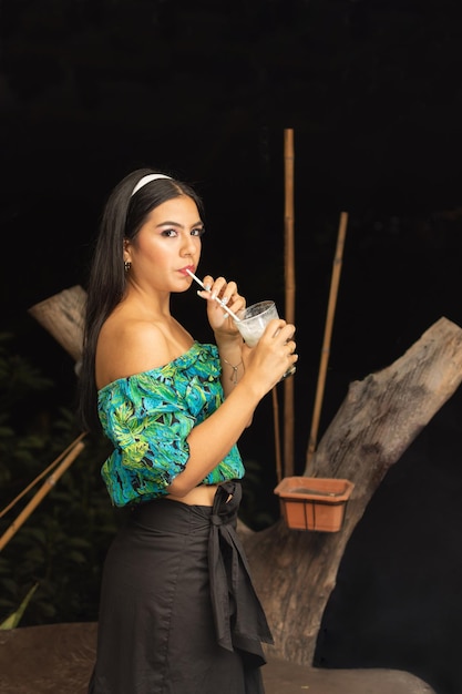 Außenporträt einer schönen jungen hispanischen Frau mit langen schwarzen Haaren, die Kokosnusslimonade trinkt und vor dunklem Hintergrund in die Kamera blickt Lifestyle Beauty Girl