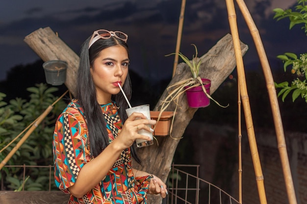 Außenporträt einer schönen jungen hispanischen Frau mit langen schwarzen Haaren, einer Sonnenbrille über dem Kopf und einem farbenfrohen Kleid, das Kokosnusslimonade trinkt und bei Sonnenuntergang in die Kamera schaut Lifestyle Beauty Girl