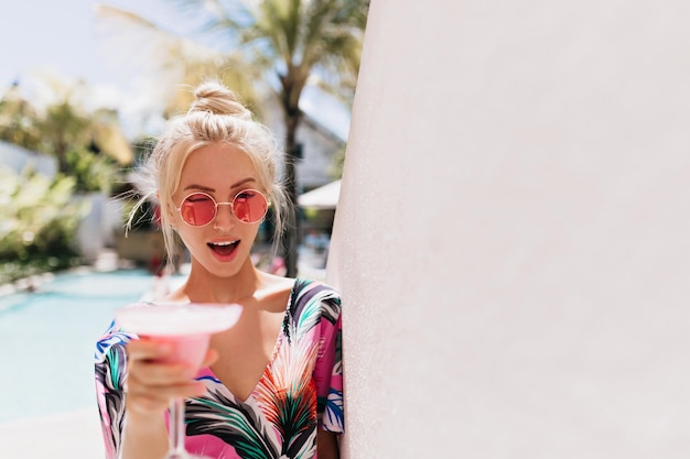 Außenporträt eines blonden, reizenden Mädchens mit rosa Sonnenbrille Foto eines wunderschönen blonden weiblichen Models, das sich im Sommerresort amüsiert und einen Cocktail trinkt