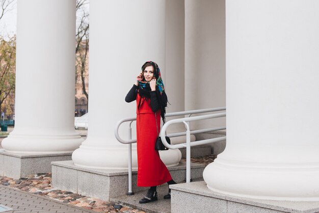 Außenporträt eines stilvollen jungen Mädchens auf der Straße einer alten Stadt in der schwarzen Kleidung und einer langen roten Weste und einem russischen Schal auf ihrem Kopf. Ein Model posiert auf der Straße einer europäischen Stadt