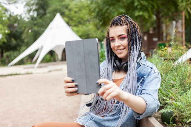 Außergewöhnliche Studentin Millennial mit afrikanischen Zöpfen mit Tablet im Freien