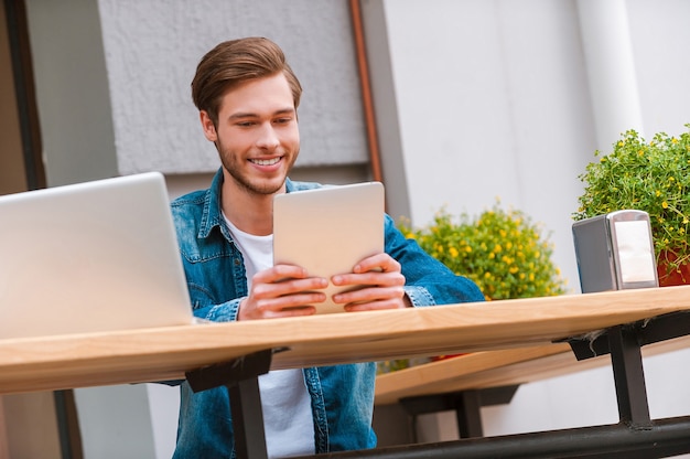 Außerhalb arbeiten. Glücklicher junger Mann, der ein digitales Tablet hält und lächelt, während er im Straßencafé sitzt?