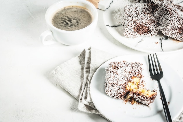 Australisches Essen Traditionelles Dessert Lamington - Stücke Keks in dunkler Schokolade mit Kokospulver-Chips bestreut Auf einer Marmorplatte weißen Tisch Mit Kaffeetasse