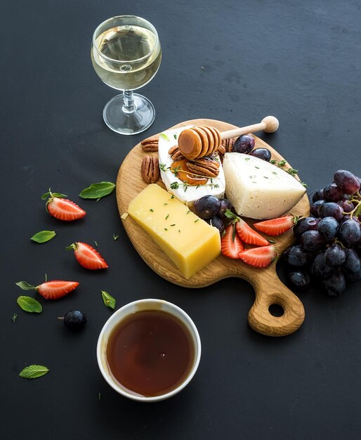 Auswahl an Käse-Vorspeisen oder Wein-Snack-Set Verschiedene Käsetrauben, Pekannüsse, Erdbeeren und Honig auf rundem Holzbrett vor schwarzem Hintergrund