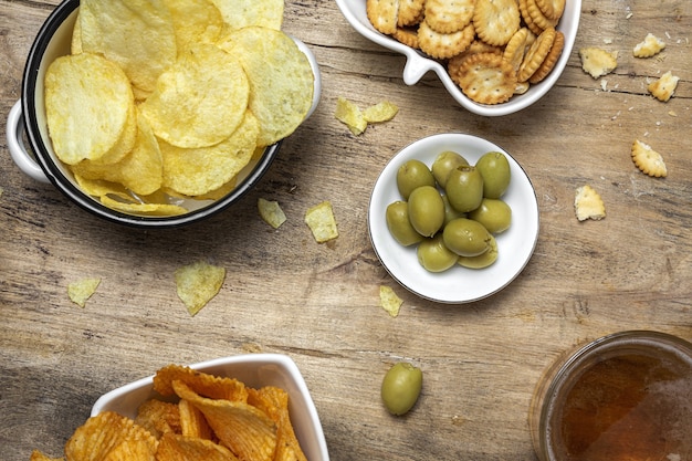 Auswahl an Snacks zu Hause mit Kartoffelchips, Bier, Crackern, grünen und schwarzen Oliven auf Holztisch