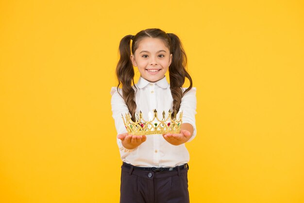 Auszeichnung für den besten Studentenpreis. Glückliches kleines Kind, das Kronenbelohnung auf gelbem Hintergrund ausgibt. Kleines Schulmädchen, das mit Juwelbelohnung lächelt. Erfolg belohnen.