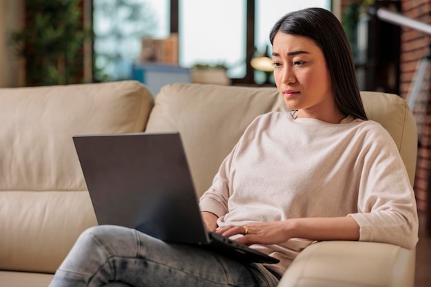 Auténtica joven asiática usando una computadora portátil, sentada en el sofá en casa, hermosa chica comprando chateando en línea en las redes sociales, viendo películas en el tiempo libre, freelance trabajando en un nuevo proyecto