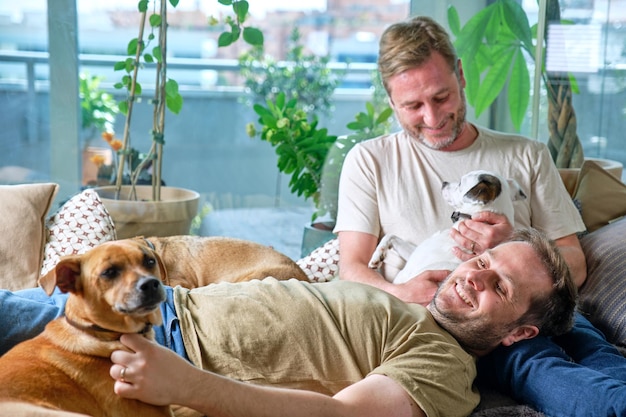 Autêntico casal gay sorrindo relaxado no sofá em casa acariciando seus três cachorros
