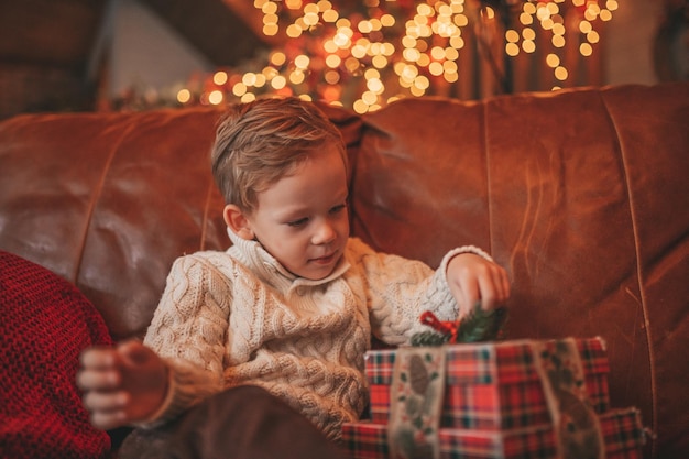 Auténtico niño feliz sincero con suéter beige de punto sentado con regalos en el albergue decorado con Navidad