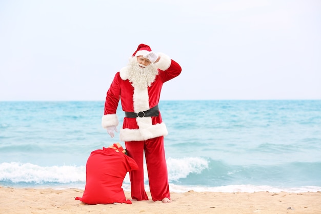 Foto auténtico papá noel con gran bolsa roja llena de regalos en la playa