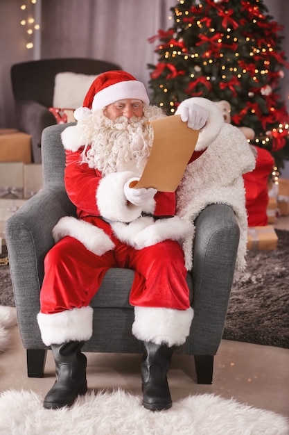 Auténtico Papá Noel con hoja de papel sentado en un sillón en la habitación decorada para Navidad