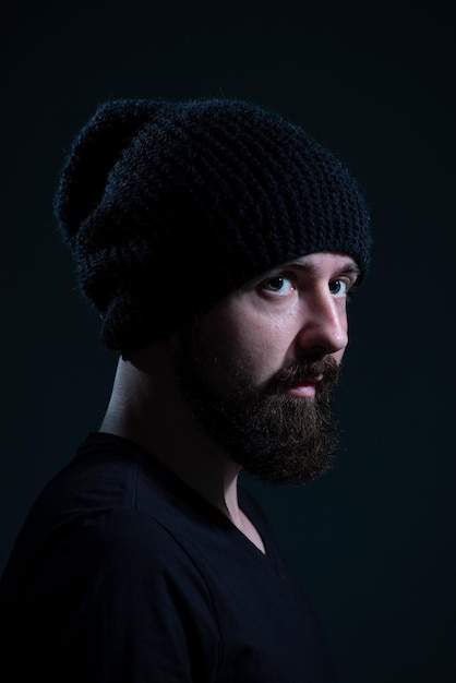 Auténtico retrato dramático de un milenario con barba en un sombrero de punto negro sobre un fondo oscuro