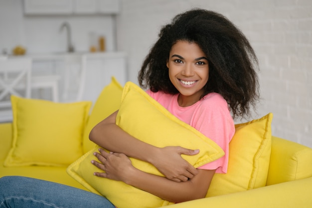 Auténtico retrato de linda chica de pelo rizado emocional relajante y sonriente en casa. Joven y atractiva mujer afroamericana abrazando la almohada sentada en un cómodo sofá amarillo en la sala de estar