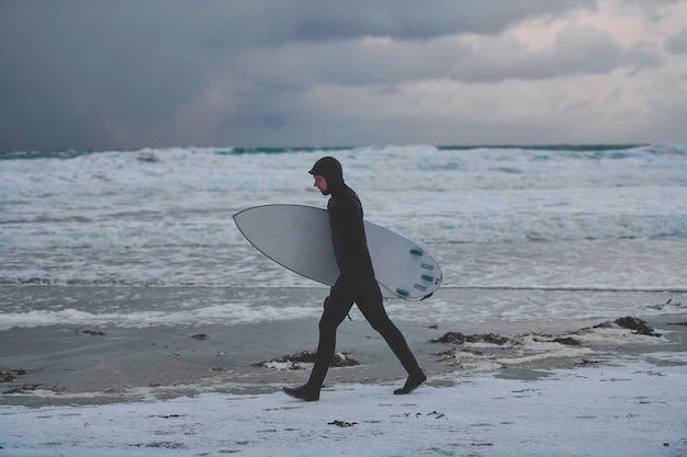 Autêntico surfista do Ártico local indo pela praia depois de surfar no mar do Norte. Litoral do mar norueguês. Esportes radicais de atividades aquáticas de inverno