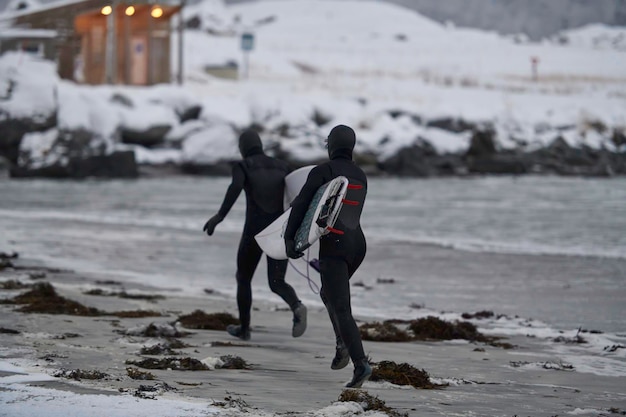 Auténticos surfistas locales del Ártico corriendo en una playa nevada después de surfear en el mar noruego. Fondo de montaña cubierto de nieve en invierno. Islas Lofoten noruegas. Costa del mar noruego. Actividad de agua de invierno