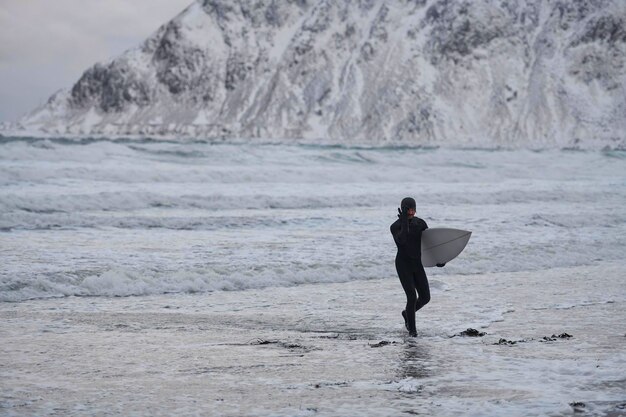 Authentischer lokaler arktischer Surfer, der nach dem Surfen in der Nordsee am Strand vorbeigeht. Schneebedeckte Berge im Hintergrund. Norwegische Meeresküste. Winterwasseraktivitäten Extremsport