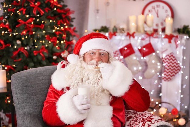 Authentischer Weihnachtsmann mit Keks und Glas Milch im Sessel im weihnachtlich dekorierten Zimmer sitzend