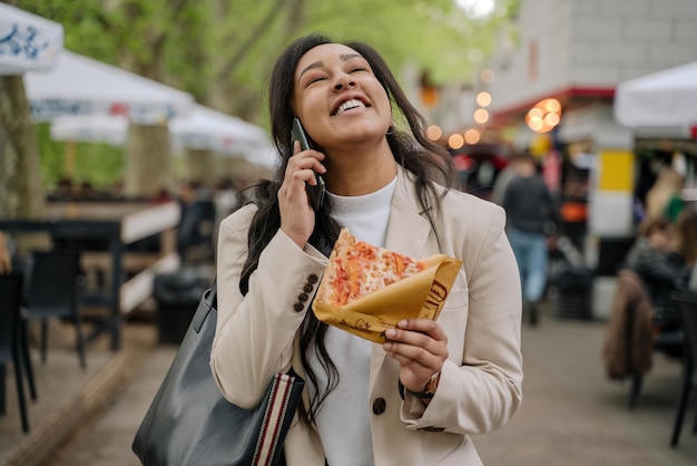 Authentisches Porträt einer schönen lächelnden Frau, die mit einem Stück Pizza telefoniert