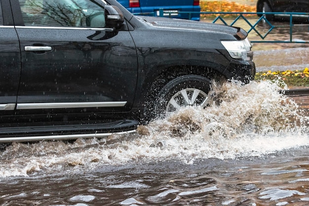 El auto está conduciendo a través de un charco bajo fuertes lluvias Salpicaduras de agua debajo de las ruedas de un auto Inundaciones y agua alta en la ciudad