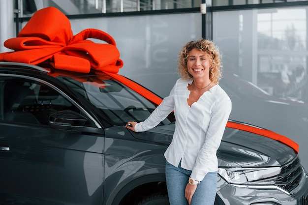 El auto está con la longitud de regalo en la parte superior La mujer con cabello rubio rizado está en autosalon