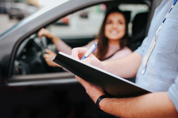 Auto instructor masculino toma examen en mujer joven. Modelo borroso sentado en el coche y tomados de la mano en el volante. Guy mantenga carpeta con papel en las manos.