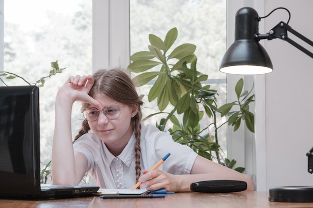 Foto auto-isolamento de colegial usando laptop para sua lição de casa. conceito de jogos, educação e escola - menina triste com laptop e em casa. rapariga com dificuldade em fazer o dever de casa online, deprimida sentada dentro de casa