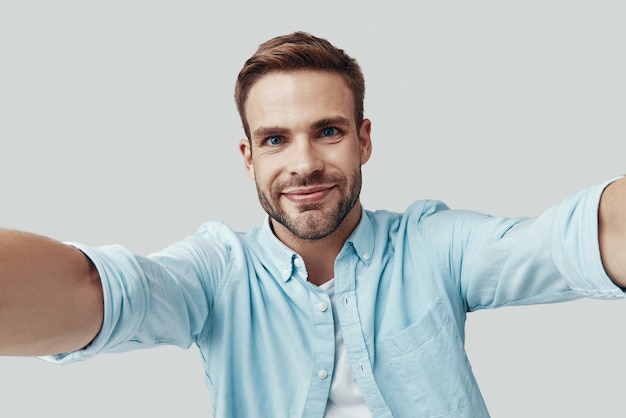 Auto-retrato de jovem bonito olhando para a câmera e sorrindo em pé contra um fundo cinza