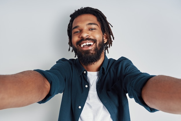 Foto auto-retrato de jovem bonito olhando para a câmera e sorrindo em pé contra uma parede cinza